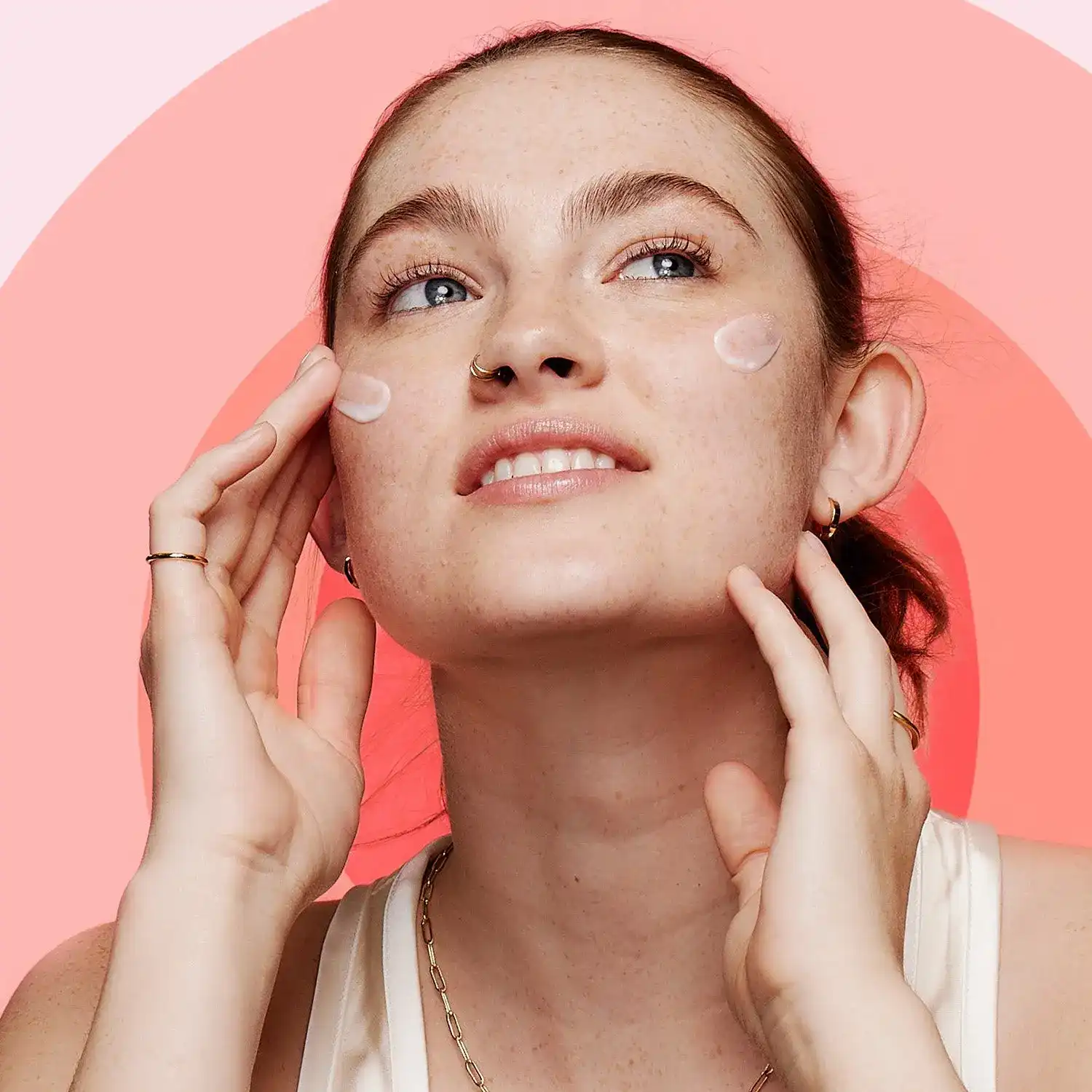A person applies cream to their cheeks, cupping their face with both hands, against a bright pink background.