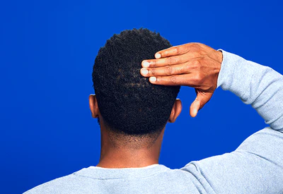 A man with dark brown hair touching the back of his head, on a blue background.