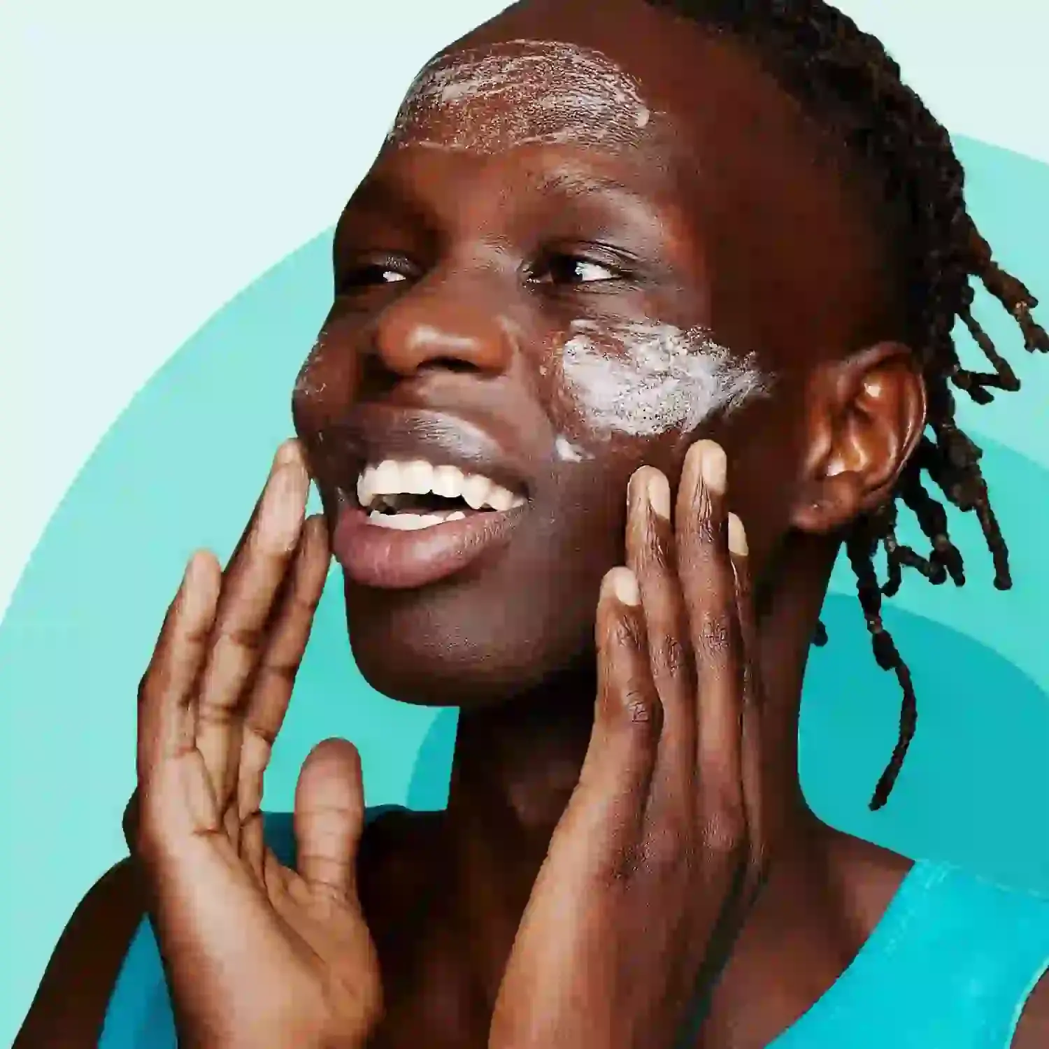 A person, smiling and applying face cream with both hands, stands against a turquoise background, exuding a sense of freshness and skincare routine.