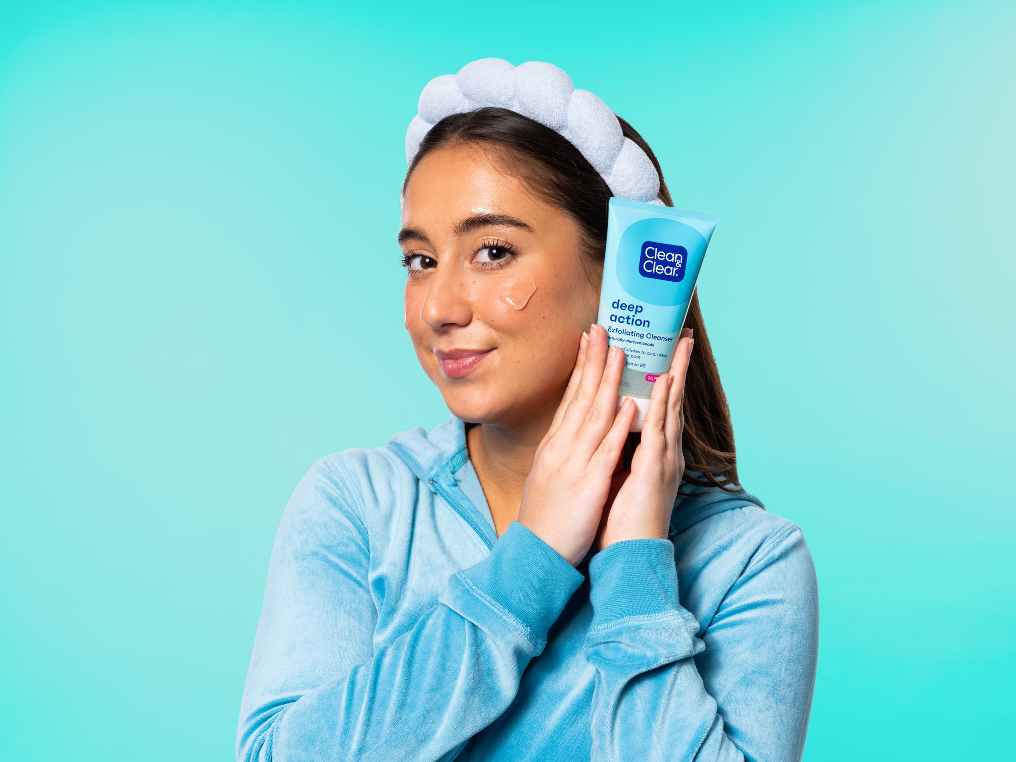 A young woman with glowing skin holding a bottle of "Deep Action" exfoliating cleanser by Clean&Clear®, standing in a blue background