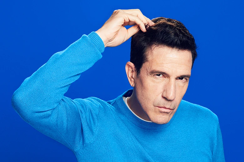 Man with brown hair and a long sleeve blue shirt touching his hair, on a blue background.