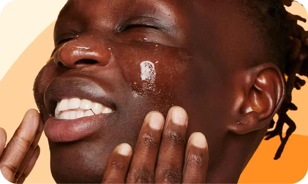 A person smiles with eyes closed, applying a glossy skincare product on their face, using their hands. The background features a gradient of light to dark orange hues.