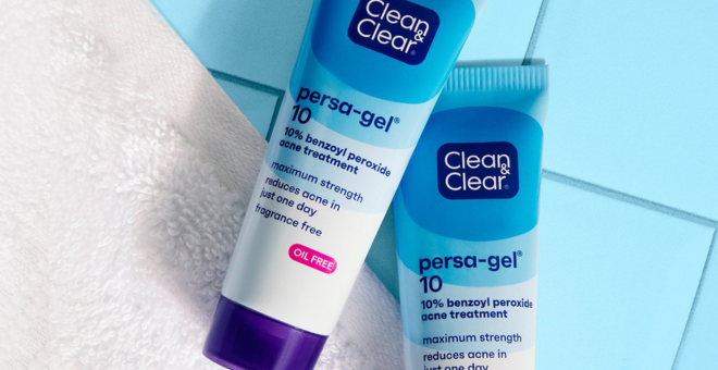 Tubes of Clean & Clear persa-gel 10 acne treatment rest on a white towel against blue tiles. Text: “10% benzoyl peroxide acne treatment, maximum strength reduces acne in just one day, fragrance free, oil free.”