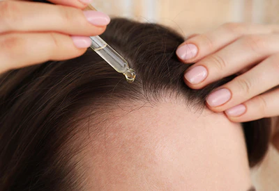 Close up of the top of a woman’s head with long brown hair applying woman’s Rogaine Topical Solution.