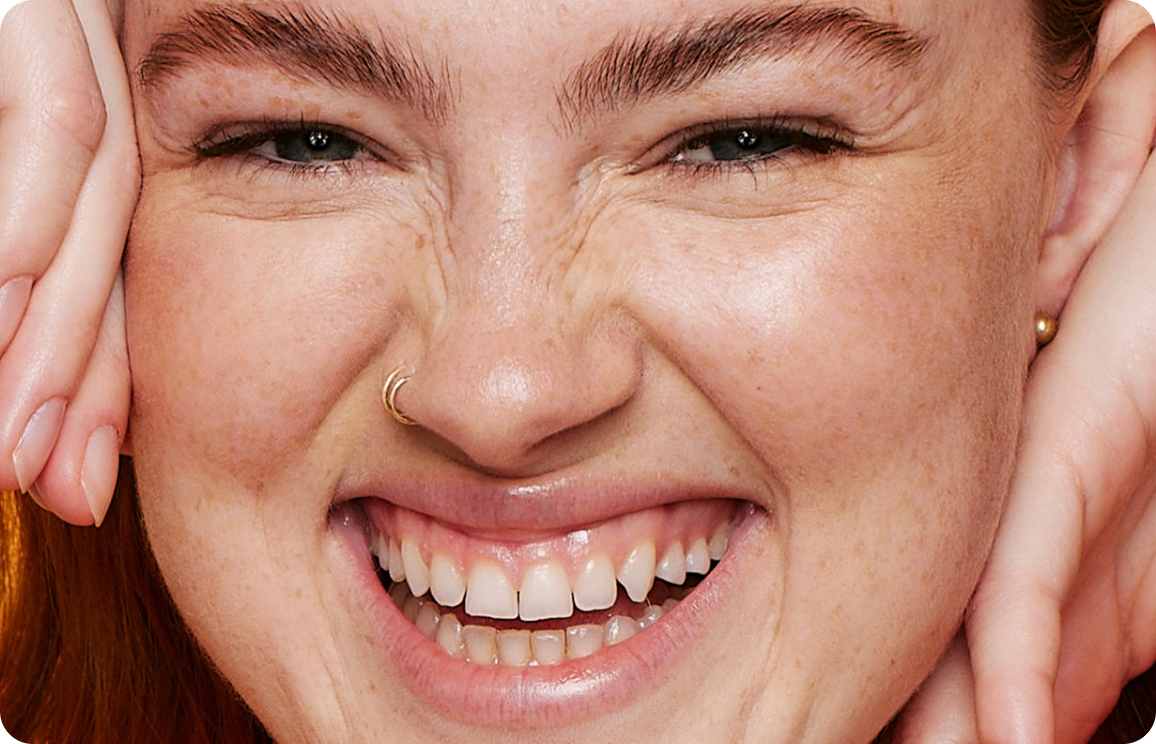 A smiling face, with a nose ring and bright eyes, framed by hands resting on cheeks, showcasing freckles and joyful expression in a close-up view.