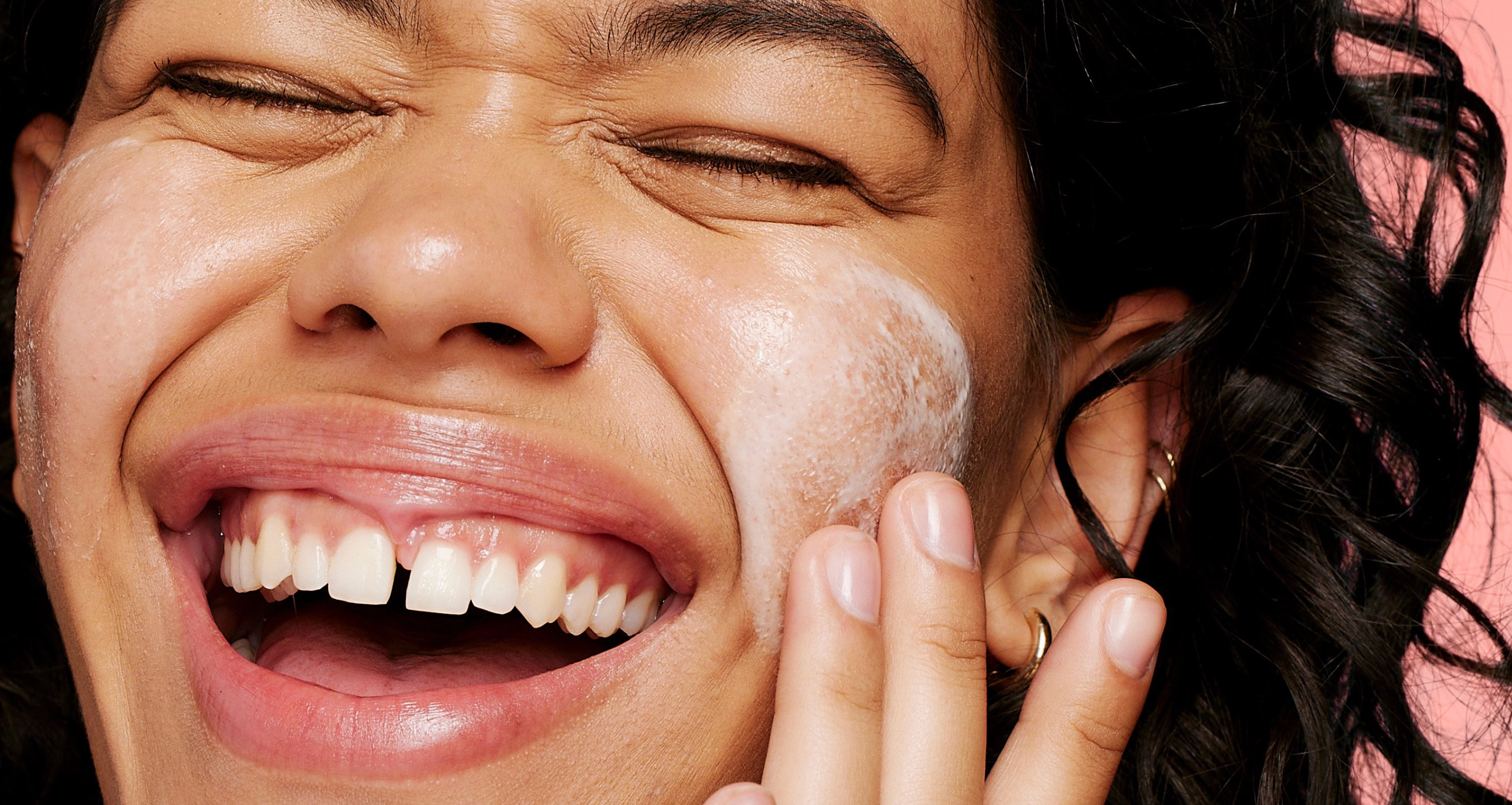 A person with curly hair smiles broadly while applying facial cleanser to their cheek against a pink background.