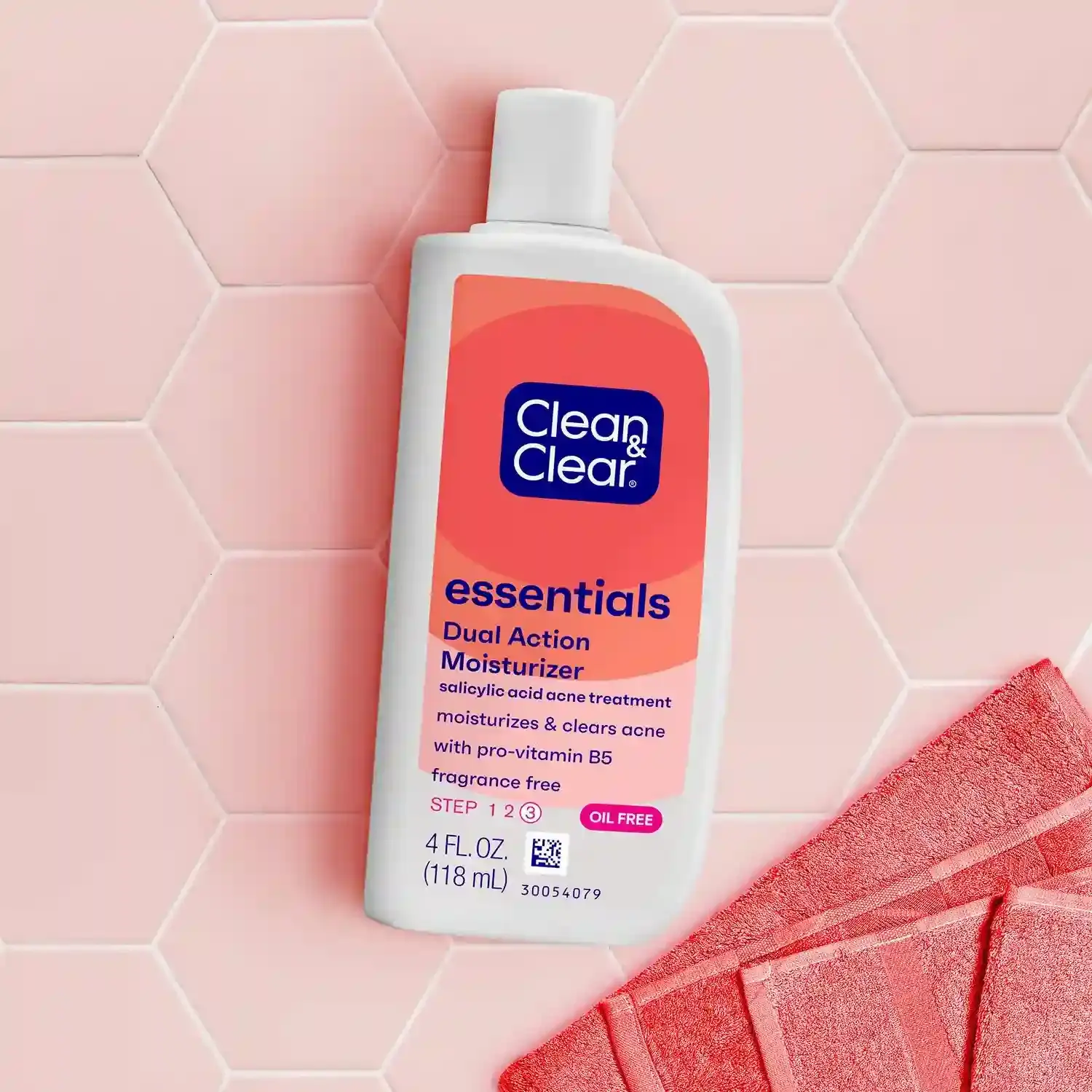 A bottle of Clean & Clear Essentials Dual Action Moisturizer lies on a pale hexagonal tiled surface, near a folded red towel. Text includes “salicylic acid acne treatment,” “fragrance free,” “4 FL. OZ. (118 mL),” and “30054079.”