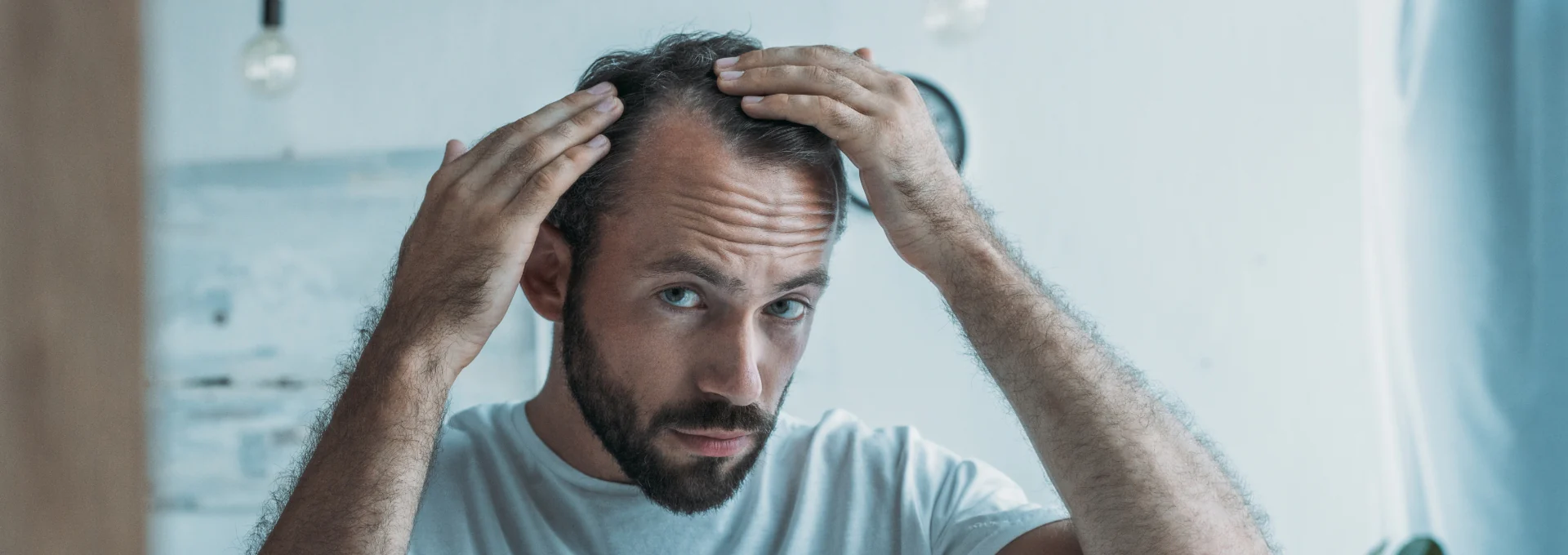 A man with light brown hair and a beard inspecting the part of his hair in a mirror.