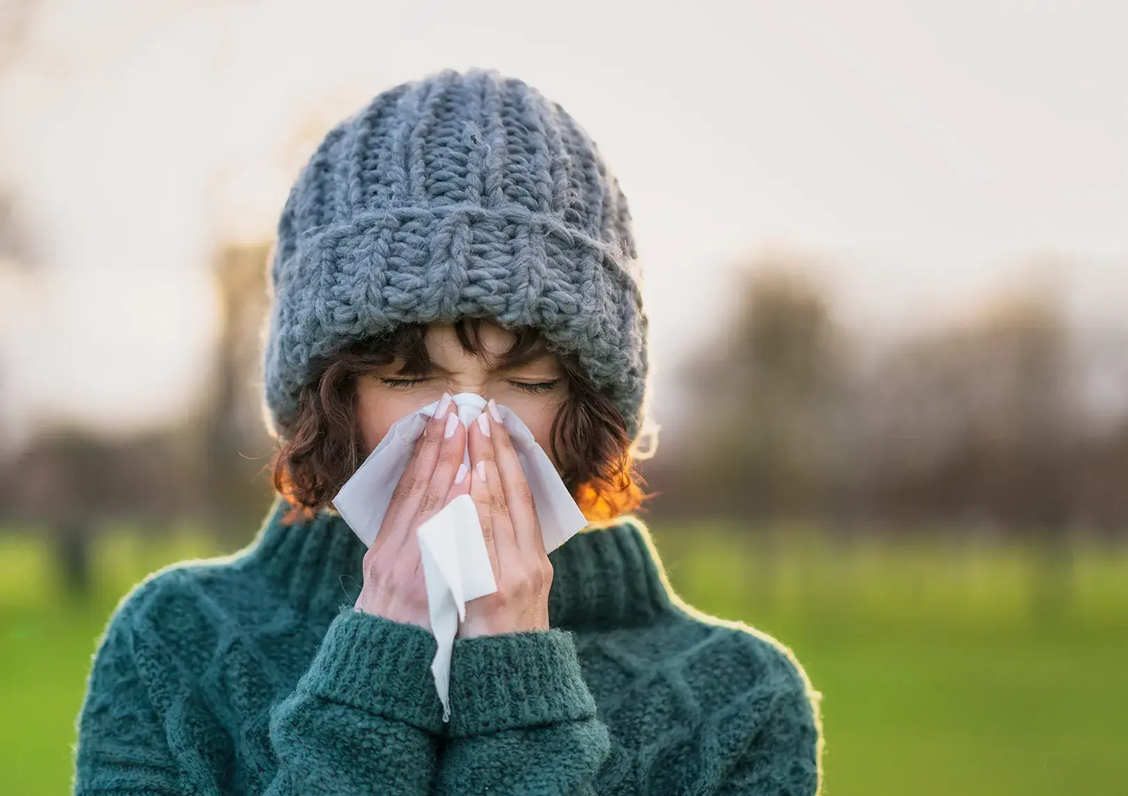 Girl wearing a gray hat and green sweater holds a tissue, visibly suffering from common cold symptoms in an outdoor setting.