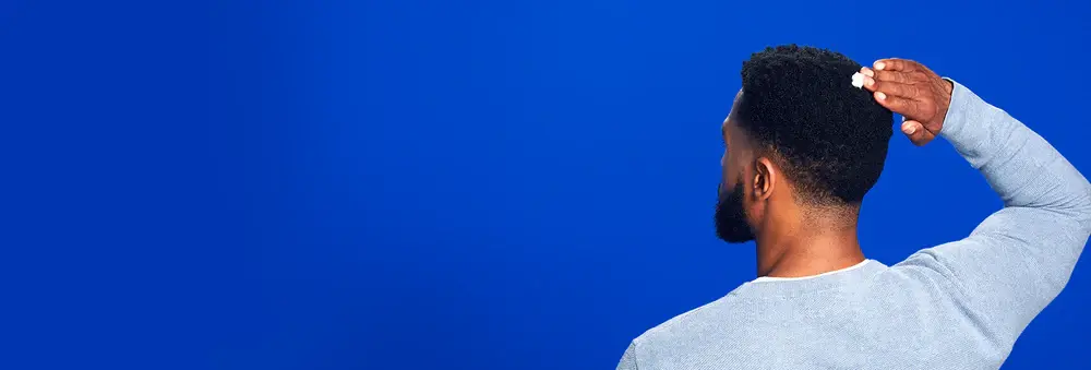 Man with dark brown hair and beard applying Men's ROGAINE® Foam to the back of his head on a blue background. Man with dark brown hair and beard applying Men's ROGAINE® Foam to the back of his head on a blue background