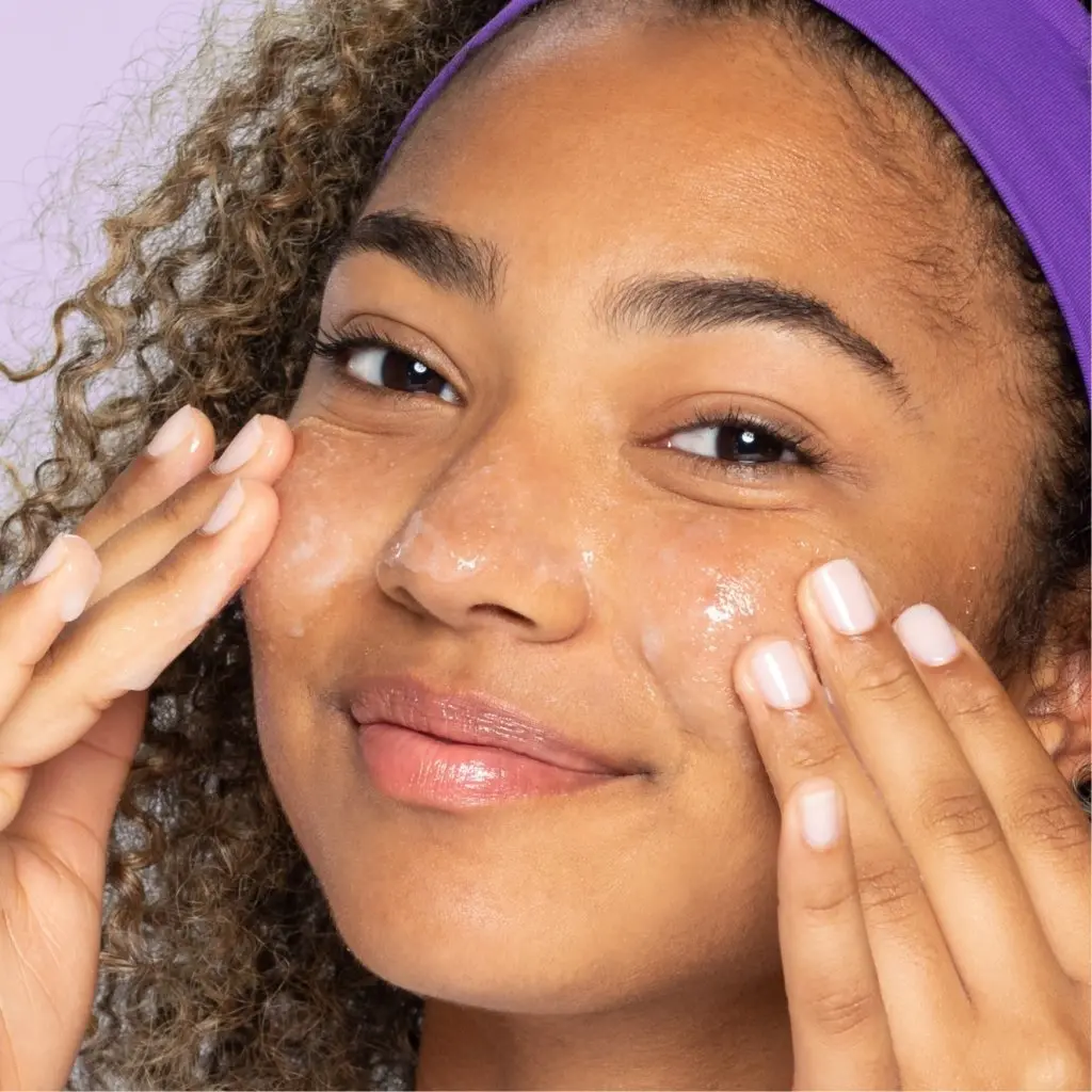 Young brown eye teen with purple headband and curls applying daily pore cleanser to cheeks