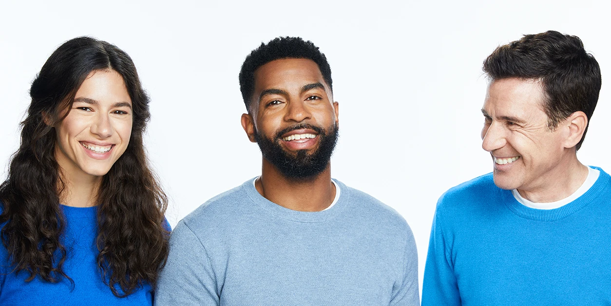 Woman with long brown hair smiling next to two men with dark hair wearing long sleeve shirts on a white background.