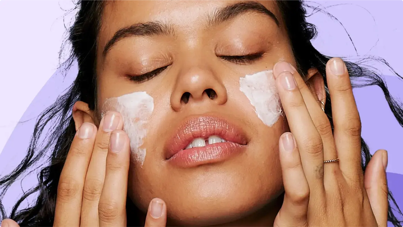 A person gently applies cream to their cheeks with closed eyes, surrounded by a soft purple background.