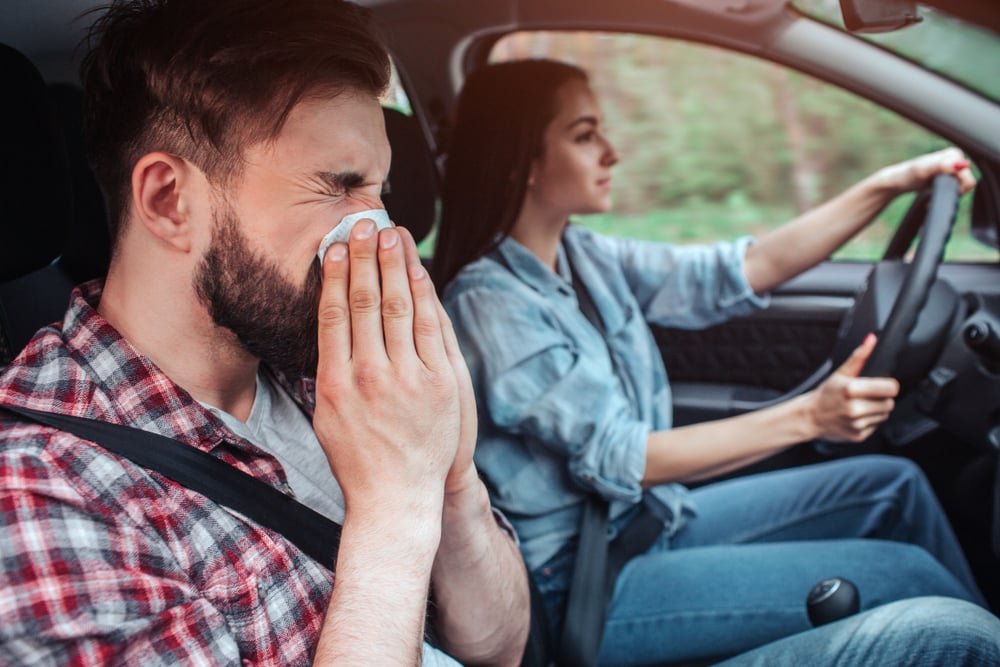 image of a woman driving a car while a man in the passenger seat blows his nose.