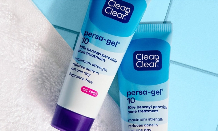 Two tubes of Clean & Clear persa-gel 10 acne treatment with purple caps rest on a blue tiled surface beside a white towel. Text: "10% benzoyl peroxide," "maximum strength," "oil free," "fragrance free."