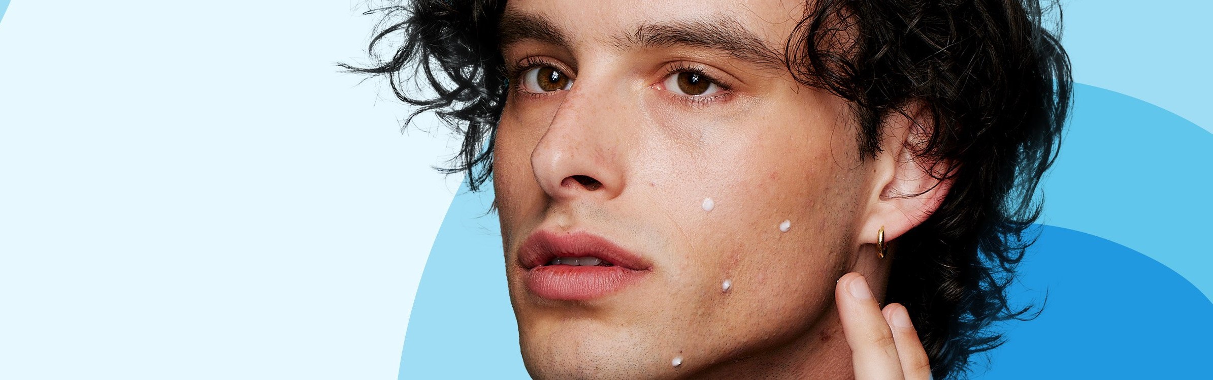 A person with curly hair applies small white dots of cream on their face, focusing intently. The background features light blue and darker blue curves.