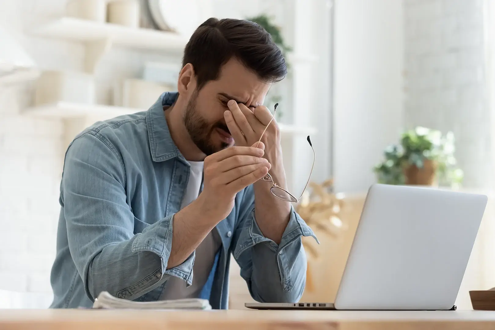 A man sitting at a desk with a laptop, indicating discomfort likely due to eye allergies.