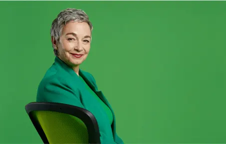 A grey haired woman in a green jacket sitting in a green chair with a green background behind her