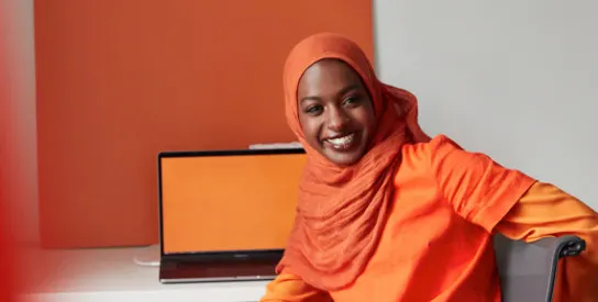 Woman in a bright orange hijab with a laptop smiling.