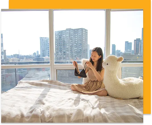 A woman is sitting on her bed next to a large stuffed animal holding clothing