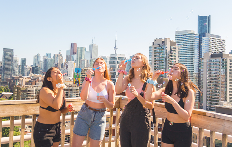 four girls standing on a balcony blowing bubbles with the city skyline in the background