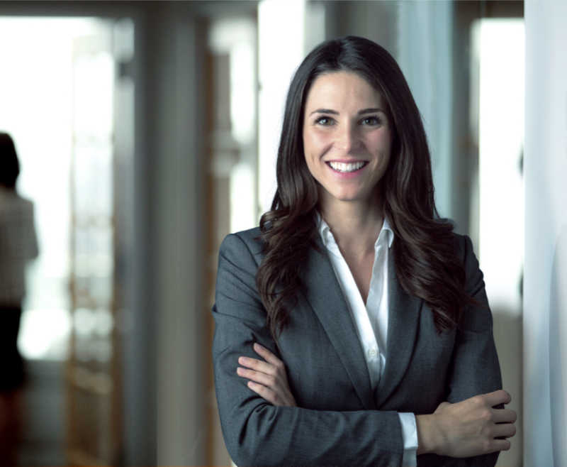 Lächelnde Geschäftsfrau mit dunklem Haar im grauen Anzug, steht mit verschränkten Armen in einem hellen Büroflur.