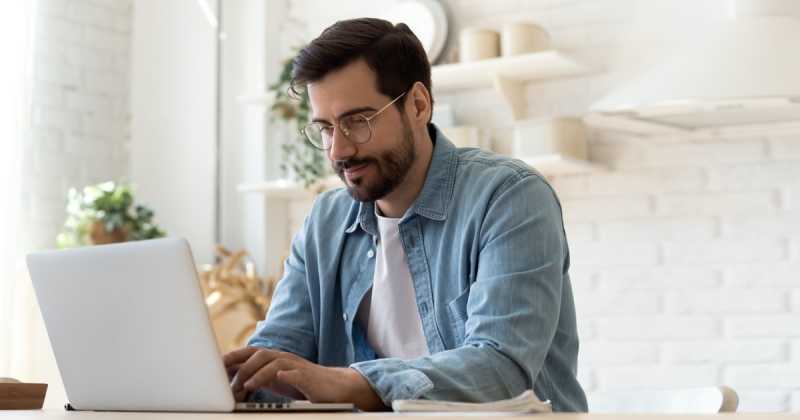 Ein Mann mit Brille und Bart, der ein Jeanshemd trägt, arbeitet in einem hellen, modernen Büro an einem Laptop.