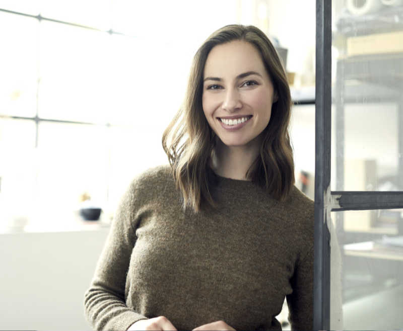 Eine Frau in einem braunen Pullover lächelt in einem hellen, modernen Büroumfeld herzlich in die Kamera.