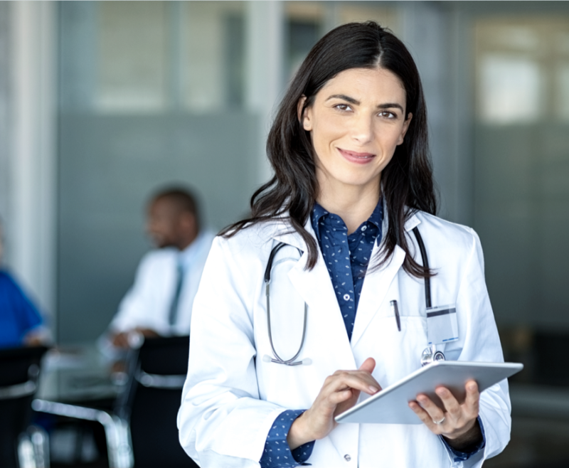 Eine Ärztin mit Stethoskop lächelt in die Kamera und hält ein Tablet in der Hand, im Hintergrund arbeiten Kollegen.