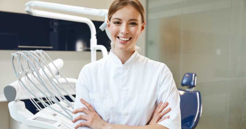 Eine lächelnde Zahnärztin im weißen Kittel steht mit verschränkten Armen in einer modernen Zahnklinik.