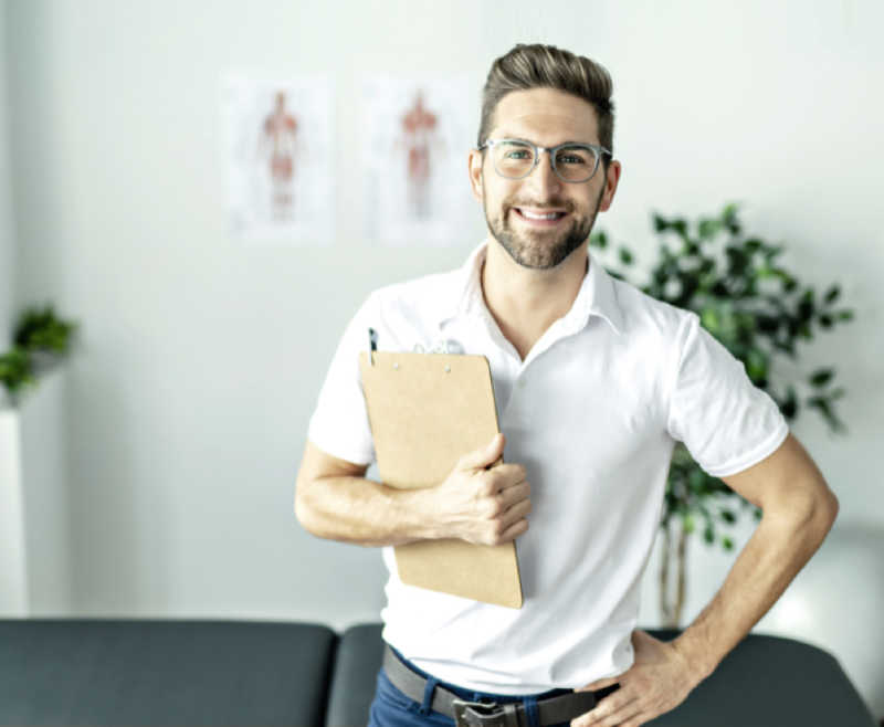Lächelnder Mann mit Brille und weißem Hemd, der ein Klemmbrett hält und in einer hellen Büroumgebung steht.
