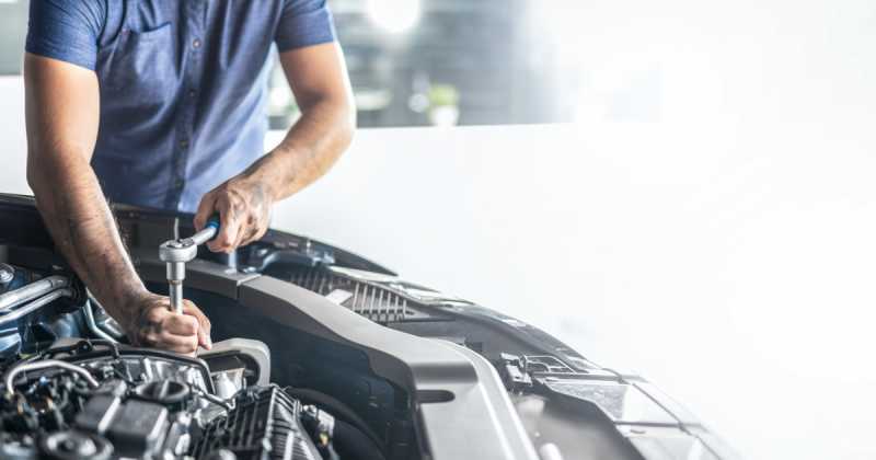 Mechaniker arbeitet mit einem Schraubenschlüssel an einem Automotor und konzentriert sich auf Details, wobei Hände und Werkzeug in einer hellen Garagenumgebung sichtbar sind.