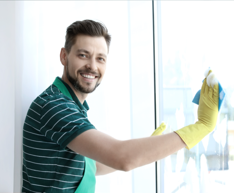 Ein lächelnder Mann mit Handschuhen putzt ein Fenster in einem hellen, sonnendurchfluteten Raum.