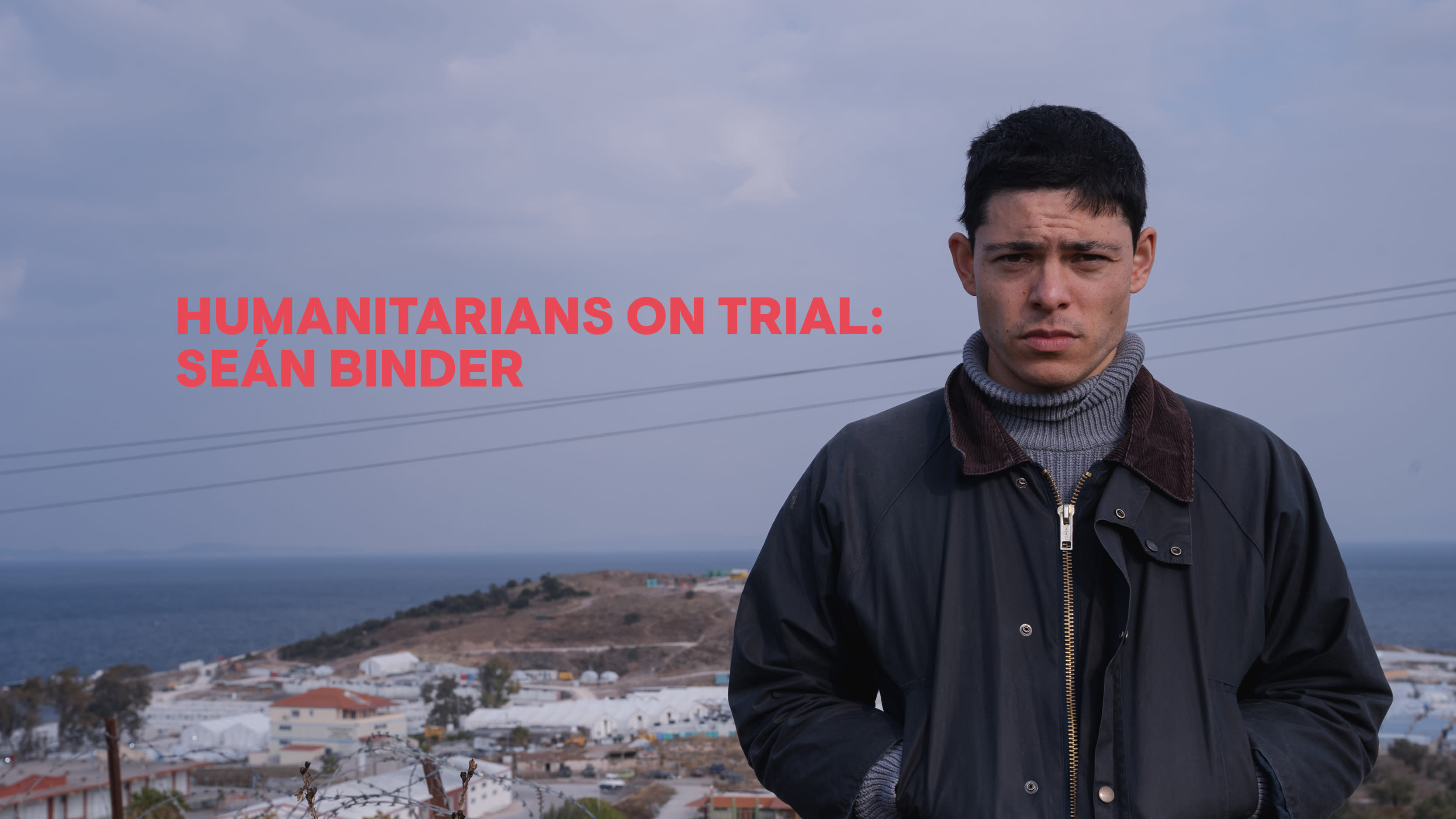 Seán faces the camera, in the background there is a view over the island of Lesvos. Text reads: 'Humanitarians on trial: Seán Binder'
