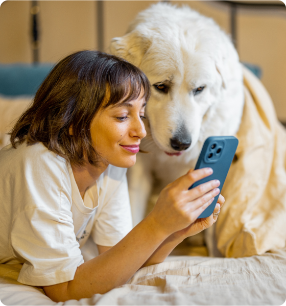A pet parent lying down on her bed with her dog. She is smiling and doing something on her smartphone.