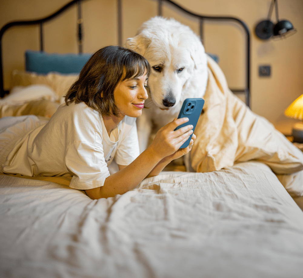 A pet parent lying down on her bed with her dog. She is smiling and doing something on her smartphone.