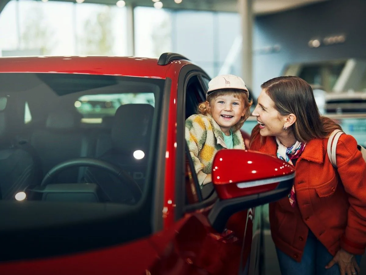 Lapsi ja aikuinen hymyilevät tutustuessaan punaiseen Volkswageniin autokaupassa