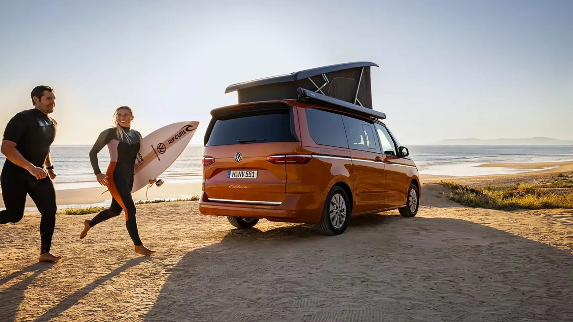 Nainen ja mies juoksevat rannalla Volkswagen California auton edestä.