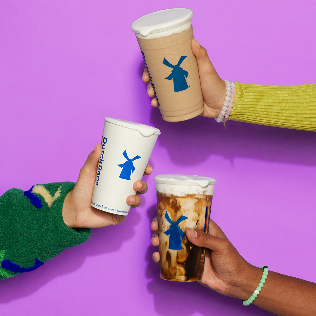 Three hands holding different Dutch Bros coffee drinks with blue windmill logos against a purple background.