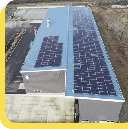 Aerial view of an industrial building with blue roof covered in solar panels, set on concrete foundation.