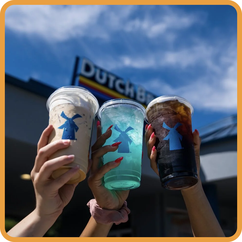 Three hands holding Dutch Bros drinks - a coffee, blue iced beverage, and dark soda - against blue sky background.
