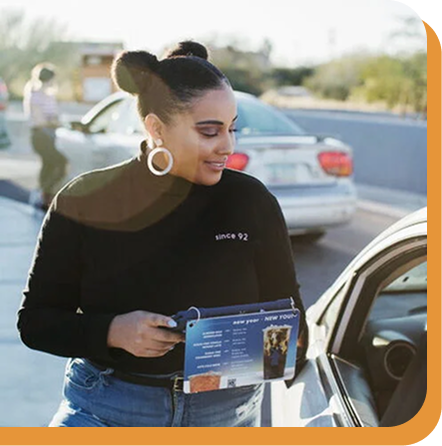 Woman with hair bun and hoop earrings wearing black top and jeans, holding an ipad next to a parked car.