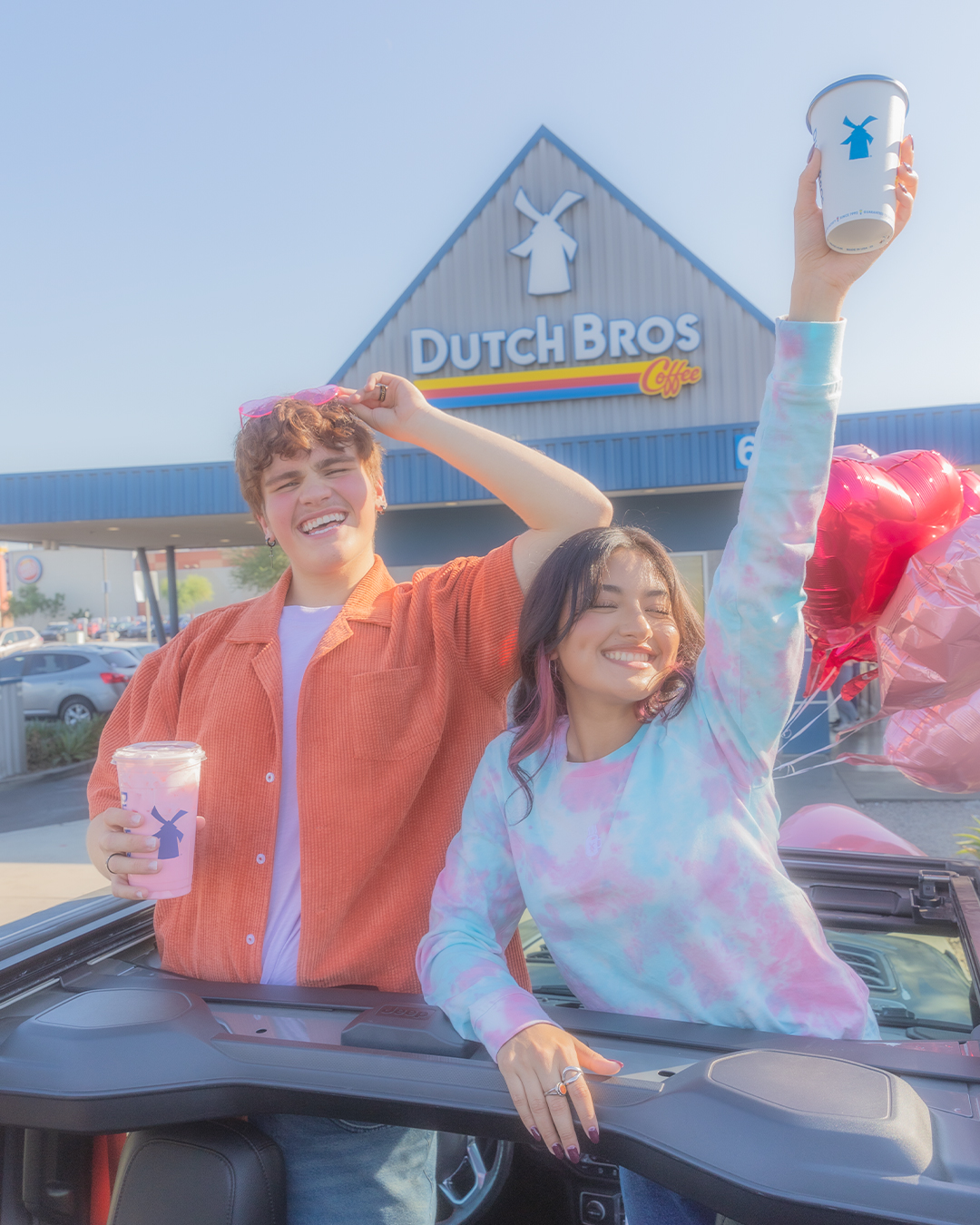 Two happy customers celebrating with Dutch Bros coffee drinks outside the store, one wearing tie-dye and holding balloons.