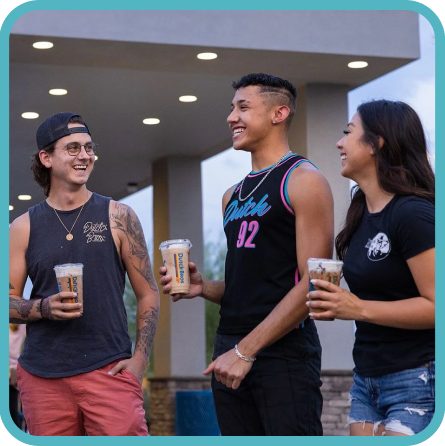 Three young people smiling and talking while holding drinks outside a building with recessed lighting.