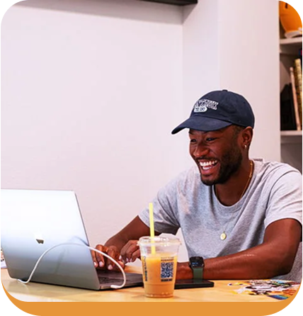 Person in blue cap smiling while working on laptop at wooden table with iced drink nearby