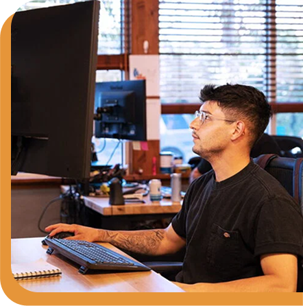Person with glasses working at computer desk with keyboard and monitor in office setting with wooden blinds.