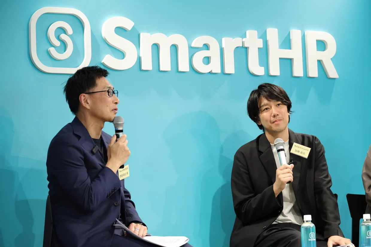 「S」ロゴと「SmartHR」の文字がある青緑色の壁を背景に、マイクを握って話す西田さんと隣の笑顔の加藤さん。