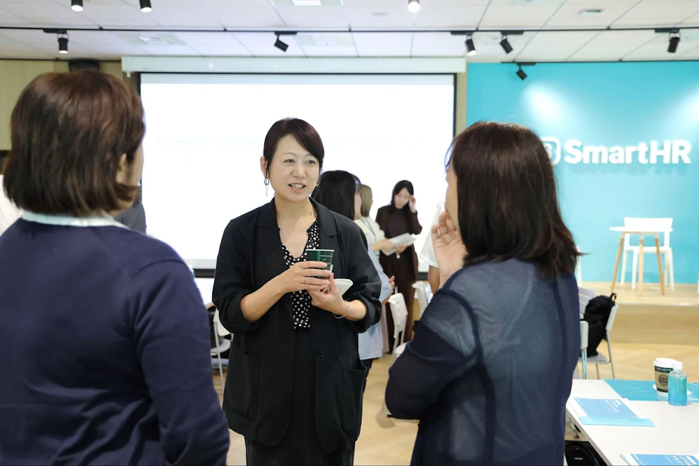 SmartHRのロゴが見える会場で、カップを手に参加者と歓談する渋谷さん