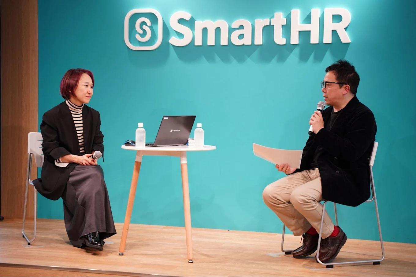 左から山崎さんと古賀さん。SmartHRのロゴがあしらわれた壁の前で対談している。2人の間には机があり、水のペットボトルとノートパソコン一台が置いてある