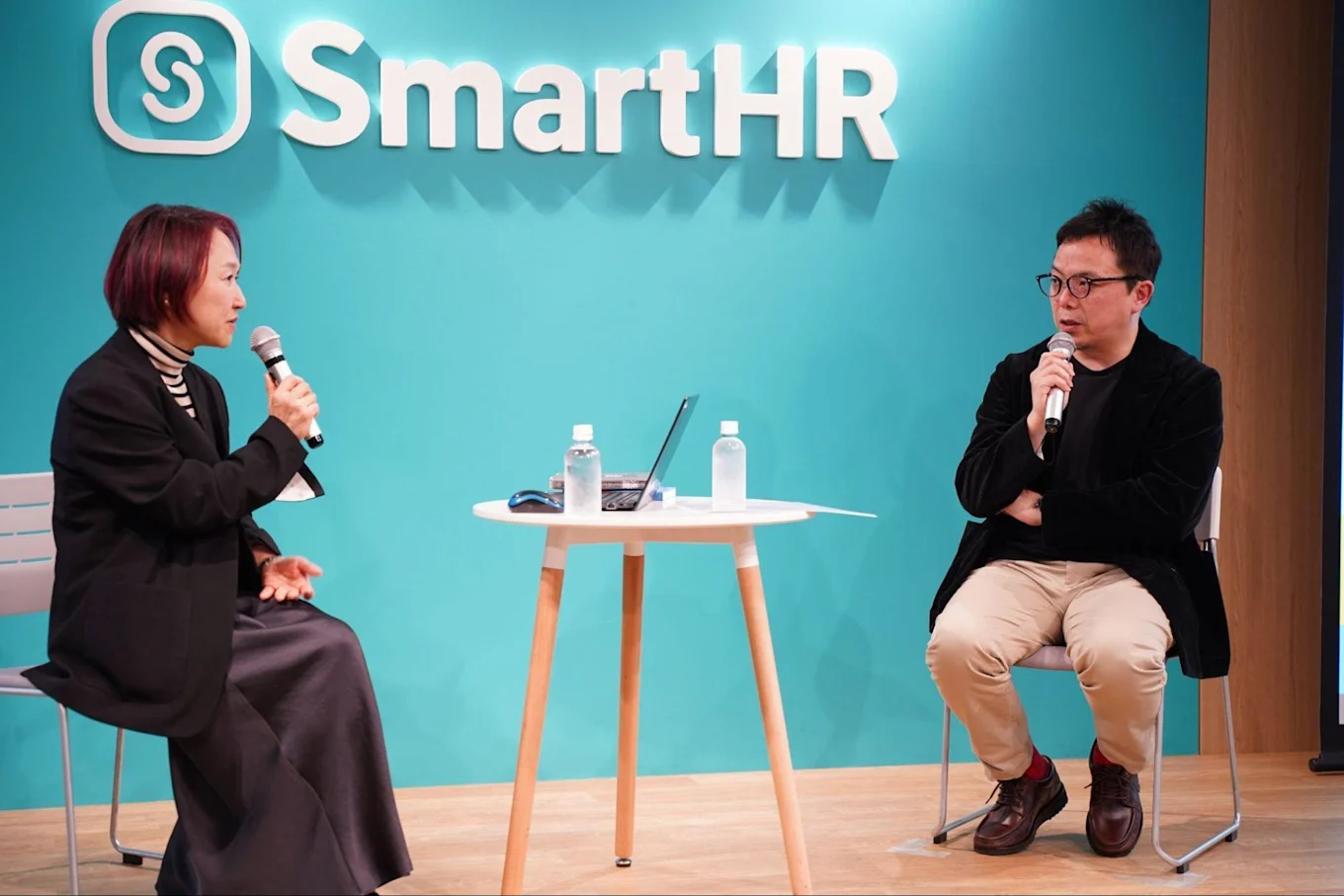 左から山崎さん、古賀さん。椅子に座り、マイクを片手にSmartHRのロゴがあしらわれた壁の前で話している